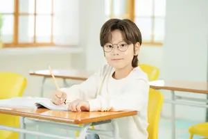 A boy with glasses sits at a desk in a classroom and writes in a notebook.