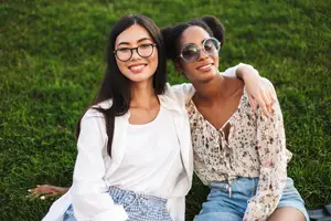 Two women sitting on a lawn, one wearing glasses and the other wearing sunglasses, both smiling and posing for a photo