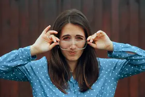 A woman is wearing a blue polka-dot shirt and holding her glasses with both hands. She is looking at something in front of her and seems to be in a room.