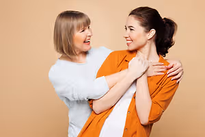 An adult woman and a young woman are smiling and posing for a photograph while holding each other's arms in a studio setting.