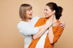An adult woman and a young woman are smiling and posing for a photograph while holding each other's arms in a studio setting.