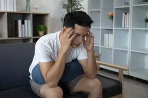 A man sitting on a couch with his eyes closed, holding his head with both hands.