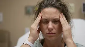 A woman in a hospital gown holding her head with both hands while looking down.