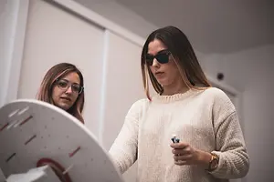 Two women, one with sunglasses and a watch, holding a pen and standing in front of a round board with a spiral pattern and lines, while the other wears glasses and a sweater, with a white wall and light fixture in the background.