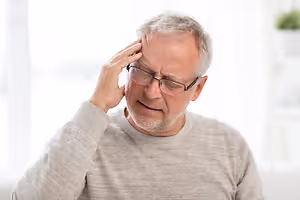 An elderly man wearing glasses and a gray long-sleeve shirt, sitting on a couch with his right hand on his head, looking to the right, possibly in a living room with a blurry view of a window, a potted plant, and a white wall behind him.