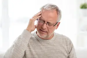 An elderly man wearing glasses and a gray long-sleeve shirt, sitting on a couch with his right hand on his head, looking to the right, possibly in a living room with a blurry view of a window, a potted plant, and a white wall behind him.