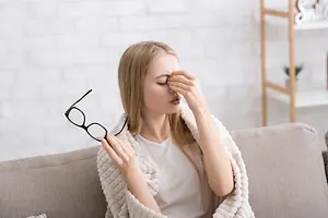 A woman sitting on a couch with a blanket on her lap, holding her nose and looking to the side, possibly in discomfort. She is wearing a white shirt and glasses. Behind her, a white brick wall with a wooden shelf on the right side. The shelf has a potted plant on top. The light is focused on the woman.