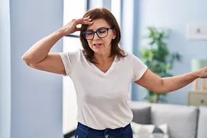 An older woman wearing glasses and a white t-shirt seems to be in pain, holding her head with one hand and her other hand raised.
