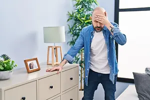 A man with a worried expression standing in a living room with his hand on his forehead.