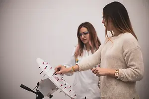 Two women in a lab, one holding a white board with red and blue pins, the other in a lab coat