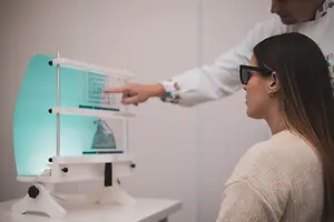 A woman wearing sunglasses looks at a monitor while a man in a white shirt points at the screen