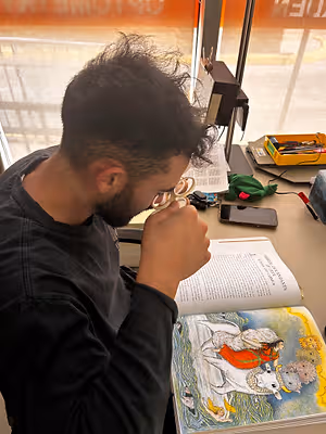 A man is sitting in front of a table with an open book and using a magnifying glass to read it.