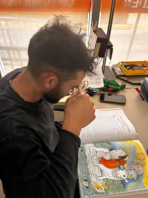 A man is sitting in front of a table with an open book and using a magnifying glass to read it.