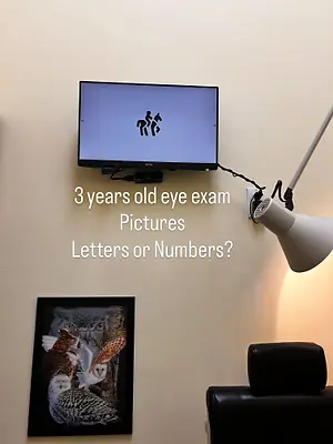 A monitor displaying a 3-year-old eye exam result with a framed picture of owls, a black chair, and a lamp.