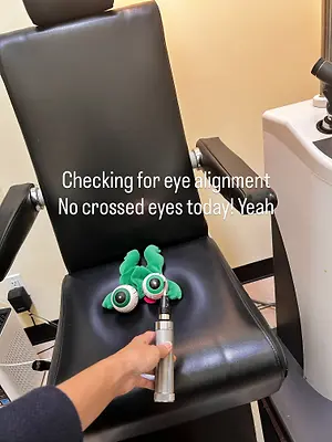 A person uses a flashlight to check the eye alignment of a green plush toy with googly eyes, saying 'Checking for eye alignment, no crossed eyes today! Yeah!'.