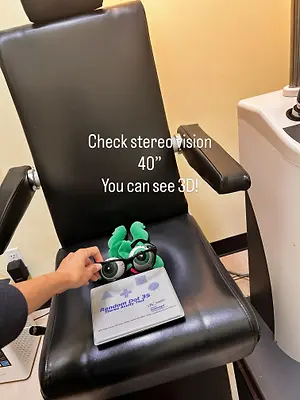 A person is sitting in a black leather chair, holding a device with the text Random Dot 3S and a toy, possibly doing a stereovision check. The device is placed on the chair, and the person is holding it with their left hand. A white device is on the right side of the chair. The room has a wooden floor and a yellow wall.