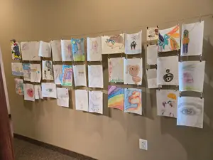 A collection of children's drawings are displayed on a wall, each drawing is unique and colorful, and they are arranged in a row with a power outlet visible on the right side of the image.