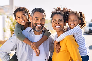 A family of three is standing in front of a white building and smiling for the camera. A man with a beard is standing between two children, and a woman with curly hair is standing behind them, smiling and looking at the camera. The man and woman are both wearing long sleeves, and the two children are wearing short sleeves.