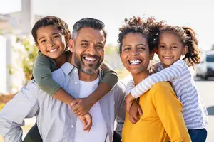 A family of three is standing in front of a white building and smiling for the camera. A man with a beard is standing between two children, and a woman with curly hair is standing behind them, smiling and looking at the camera. The man and woman are both wearing long sleeves, and the two children are wearing short sleeves.