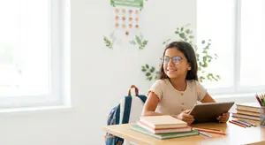 A girl with glasses sitting at a desk and smiling at a tablet