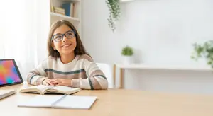 A young girl is sitting at a table, smiling, with a book, tablet, and notebook in front of her.