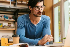 A man sitting at a desk in an office looking at a laptop with a cup of coffee beside him