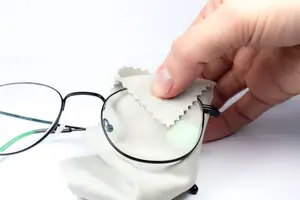 A person's hand cleaning a pair of eyeglasses with a white cloth on a white background