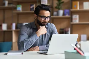 A man is sitting at a desk in a study room, working on a laptop, wearing glasses and a beard, and holding a pen in his left hand while resting his right hand on the laptop. Behind him, a wooden shelf holds books, and a potted plant sits on the shelf to his right.