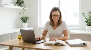 A woman sits at a desk with a laptop and book in front of her, smiling