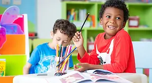 Two young children in a classroom setting, one holding a pencil and the other holding a pencil case with colored pencils. Behind them, there is a bookshelf with colorful books and a butterfly decoration on the wall.