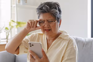 An older woman with gray hair, wearing glasses, is sitting on a couch and holding a cell phone. She is trying to see something on the phone and has a slightly open mouth.