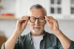 An older man holding up a pair of black glasses to his face, looking through them.