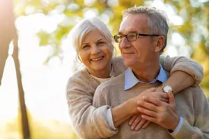 A senior couple standing close to each other and smiling, with the woman's arm around the man's shoulder, in an outdoor setting with trees and sunlight.