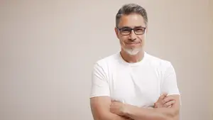 A middle-aged man with a white beard and mustache, wearing glasses and a white t-shirt, smiling and standing against a beige background.