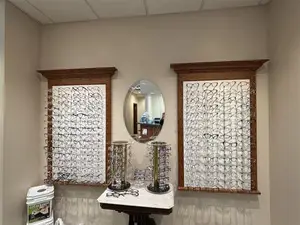 Inside a room with two framed displays of eyeglasses and a round mirror on the wall, along with a table with eyeglasses stands and a white trash bin with a label 'Got Glasses?'