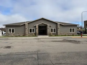 A gray building with a black front door, glass windows, and a grassy area is in an open parking lot with a streetlight and a fire hydrant on the side.