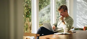 A man sitting on a chair and working on a laptop in a room with a window in the background.