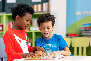 Two young boys in a classroom, one reading a book while the other smiles at him.