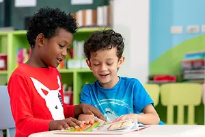 Two young boys in a classroom, one reading a book while the other smiles at him.