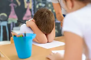A child hiding his face while sitting on a chair in a classroom