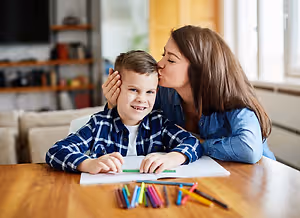 A young boy with a colorful shirt is sitting on a chair and drawing on a piece of paper with a pencil while a woman is kissing his forehead. 