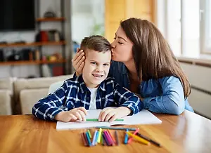 A young boy with a colorful shirt is sitting on a chair and drawing on a piece of paper with a pencil while a woman is kissing his forehead.