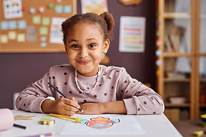 A young girl is sitting at a table in a classroom, smiling and holding a pencil while drawing on a piece of paper