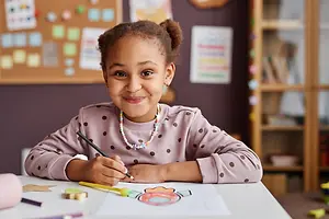 A young girl is sitting at a table in a classroom, smiling and holding a pencil while drawing on a piece of paper