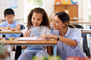 A woman with curly hair sits next to a young girl with a ponytail and they are both looking at a book on a desk in a classroom.