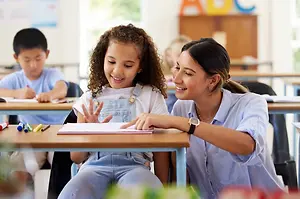 A woman with curly hair sits next to a young girl with a ponytail and they are both looking at a book on a desk in a classroom.