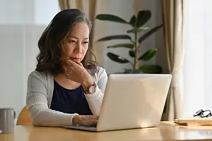 An older woman wearing a white cardigan and a watch is sitting on a chair in front of a desk, typing on a laptop and seemingly thinking about something.