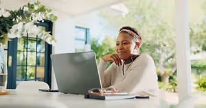 An adult woman is sitting on a chair and working on her laptop while wearing earphones on a bright sunny day