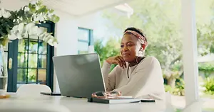 An adult woman is sitting on a chair and working on her laptop while wearing earphones on a bright sunny day