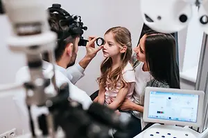 A young girl with her mother is being examined by a doctor in a room with an eye examination machine.